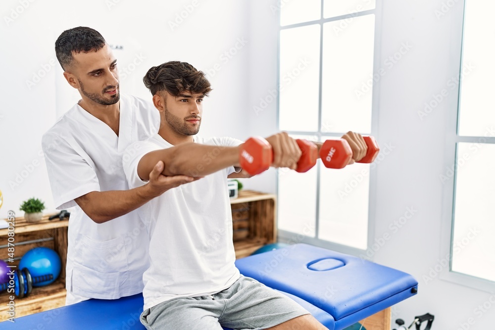Obraz premium Two hispanic men physiotherapist and patient having rehab session using dumbbells at clinic