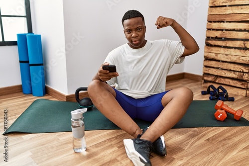 Young african man sitting on training mat at the gym using smartphone strong person showing arm muscle  confident and proud of power