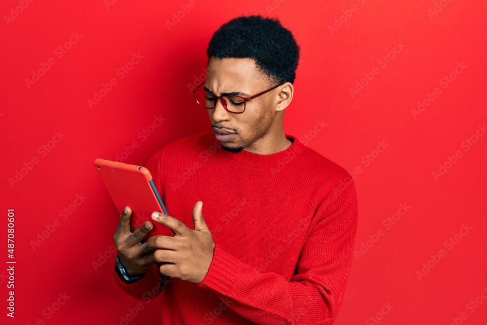 Young african american man using touchpad device thinking attitude and sober expression looking self confident Young african american man using touchpad device thinking attitude and sober expression looking self confident