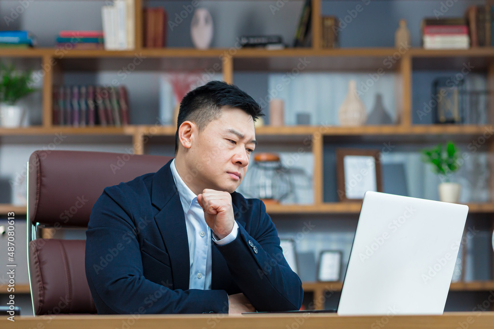 Pensive and anxious businessman, in a classic office, works at a desk with a laptop, an Asian makes important decisions, chooses a business strategy