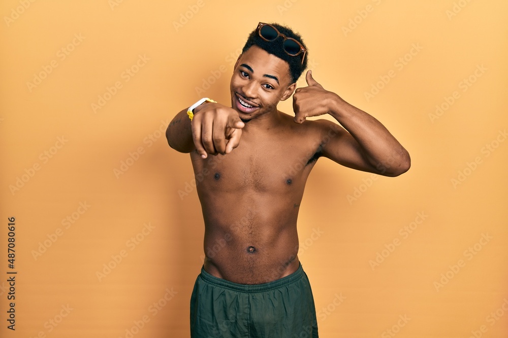 Young african american man wearing swimwear and swimmer glasses smiling doing talking on the telephone gesture and pointing to you. call me.
