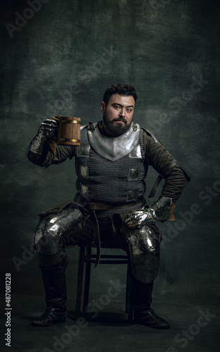 Happy medieval warrior or knight with dirty wounded face holding big mug of beer isolated over dark vintage background. Comparison of eras, history