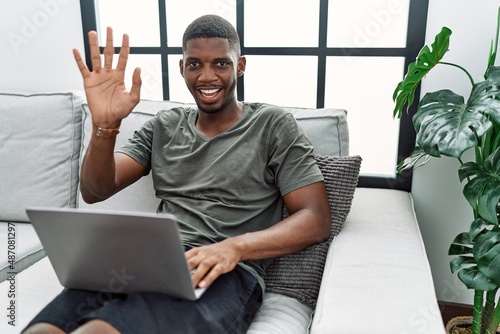 Young african american man using laptop at home sitting on the sofa waiving saying hello happy and smiling, friendly welcome gesture photo