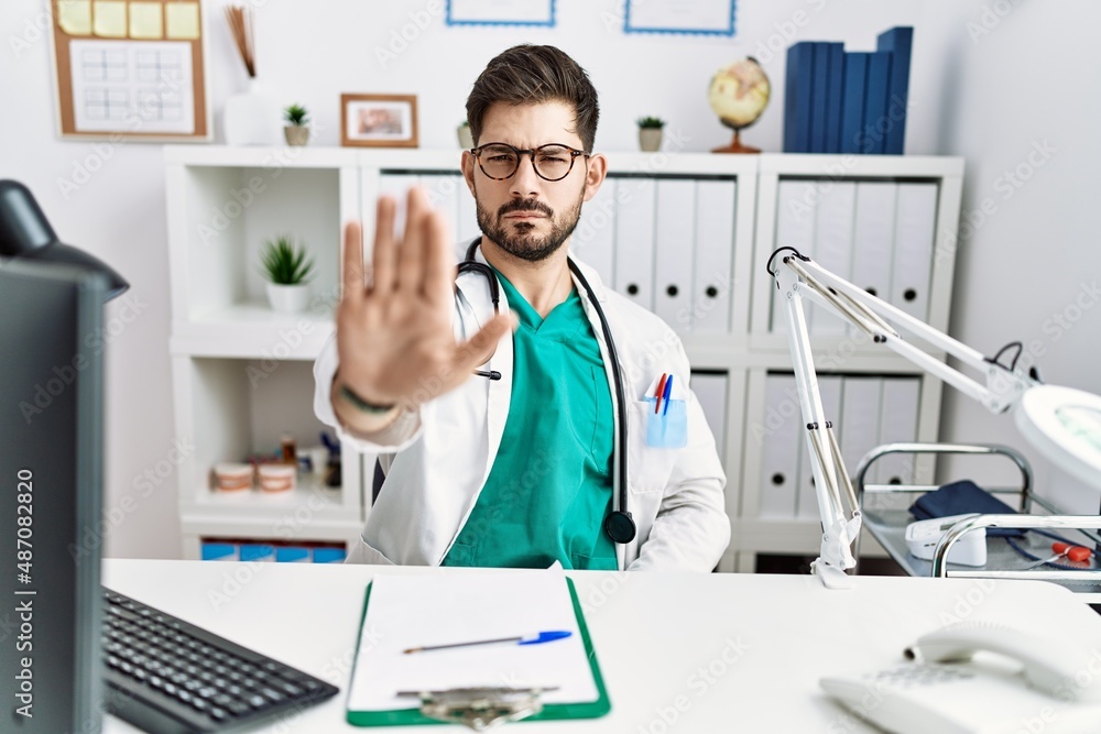 Young man with beard wearing doctor uniform and stethoscope at the clinic doing stop sing with palm of the hand. warning expression with negative and serious gesture on the face. Young man with beard wearing doctor uniform and stethoscope at the clinic doing stop sing with palm of the hand. warning expression with negative and serious gesture on the face.