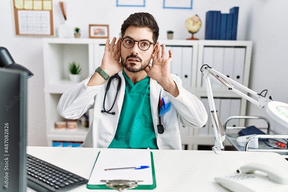 Young man with beard wearing doctor uniform and stethoscope at the clinic trying to hear both hands on ear gesture, curious for gossip. hearing problem, deaf Young man with beard wearing doctor uniform and stethoscope at the clinic trying to hear both hands on ear gesture, curious for gossip. hearing problem, deaf
