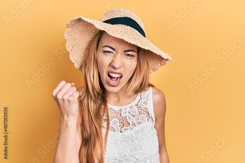Young caucasian woman wearing summer hat angry and mad raising fist frustrated and furious while shouting with anger. rage and aggressive concept.