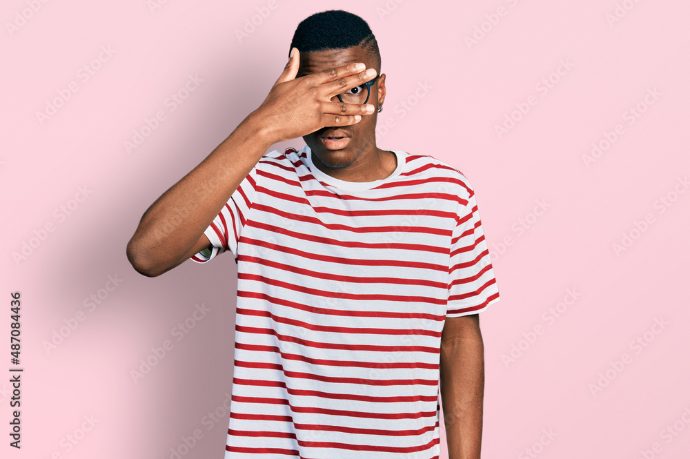Young african american man wearing casual t shirt and glasses peeking in shock covering face and eyes with hand, looking through fingers with embarrassed expression. Young african american man wearing casual t shirt and glasses peeking in shock covering face and eyes with hand, looking through fingers with embarrassed expression.