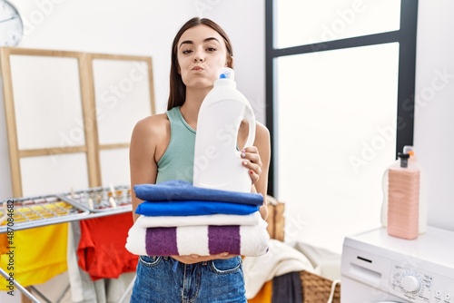 Young hispanic woman doing laundry holding detergent bottle and folded clothes puffing cheeks with funny face. mouth inflated with air, catching air.