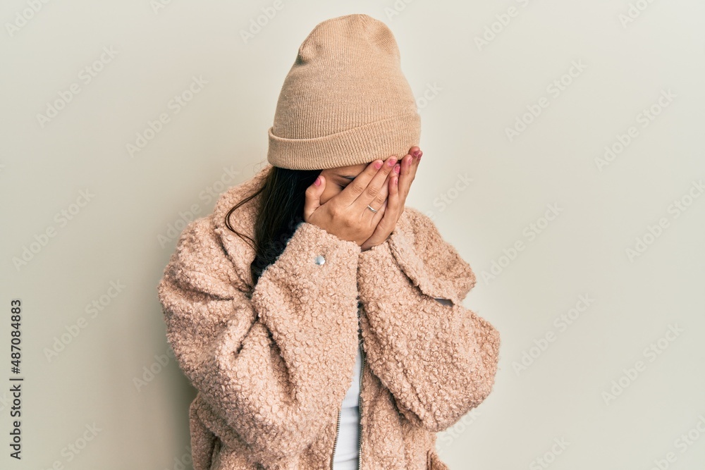 Young hispanic woman wearing wool sweater and winter hat with sad expression covering face with hands while crying. depression concept.