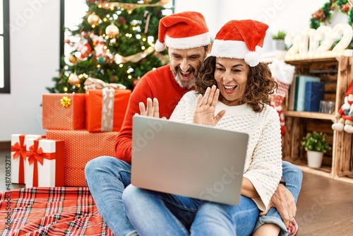 Middle age hispanic couple wearing christmas hat. Sitting on the floor having video call using laptop at home.