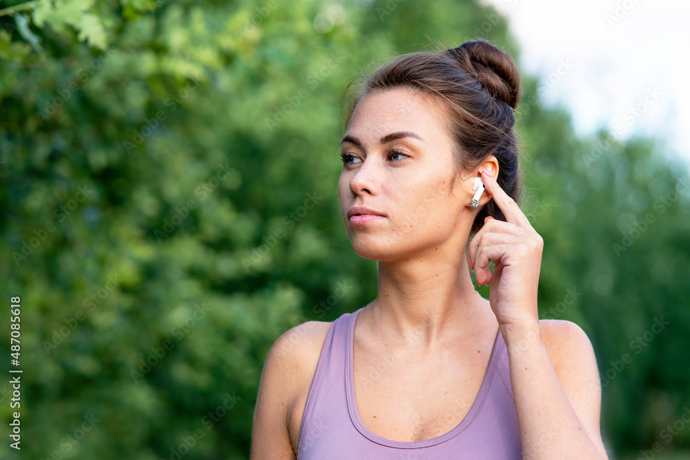 Close-up portrait of a beautiful young, pensive woman talking through bluetooth headphones while walking in the park. Serious girl, listens to music in bluetooth wireless headphones, in summer. Close-up portrait of a beautiful young, pensive woman talking through bluetooth headphones while walking in the park. Serious girl, listens to music in bluetooth wireless headphones, in summer.