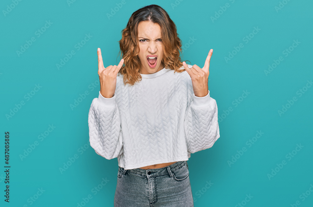Beautiful young caucasian woman wearing casual winter sweater shouting with crazy expression doing rock symbol with hands up. music star. heavy concept. Beautiful young caucasian woman wearing casual winter sweater shouting with crazy expression doing rock symbol with hands up. music star. heavy concept.