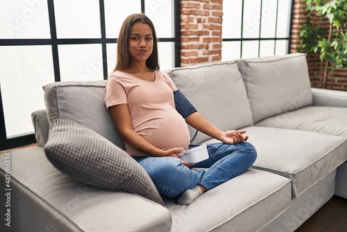 Young pregnant woman using blood pressure monitor sitting on the sofa puffing cheeks with funny face. mouth inflated with air, catching air.