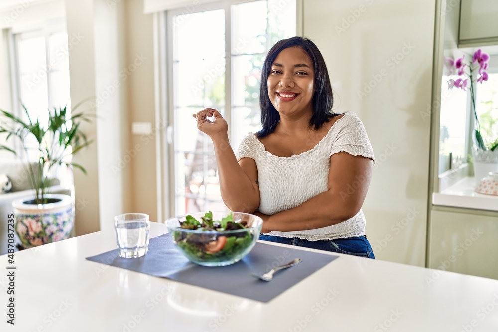 Young hispanic woman eating healthy salad at home with a big smile on face, pointing with hand and finger to the side looking at the camera. Young hispanic woman eating healthy salad at home with a big smile on face, pointing with hand and finger to the side looking at the camera.