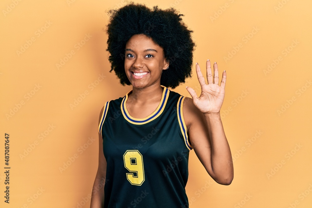 Young african american woman wearing basketball uniform showing and pointing up with fingers number five while smiling confident and happy.