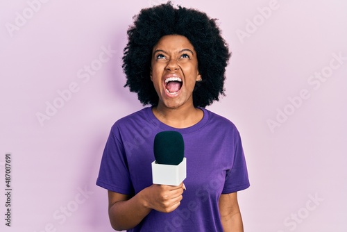 Young african american woman holding reporter microphone angry and mad screaming frustrated and furious, shouting with anger looking up.