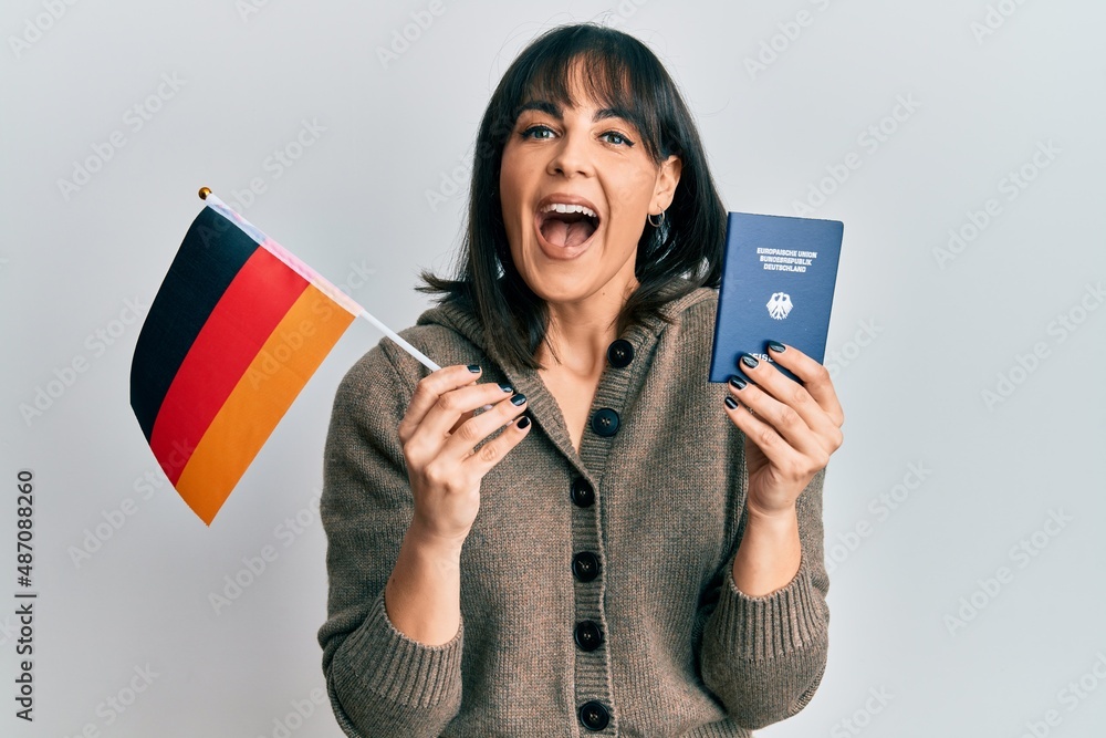 Naklejka premium Young hispanic woman holding germany flag and passport celebrating crazy and amazed for success with open eyes screaming excited.