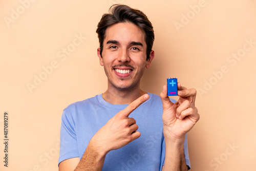 Young caucasian man holding a batterie isolated on beige background smiling and pointing aside, showing something at blank space.