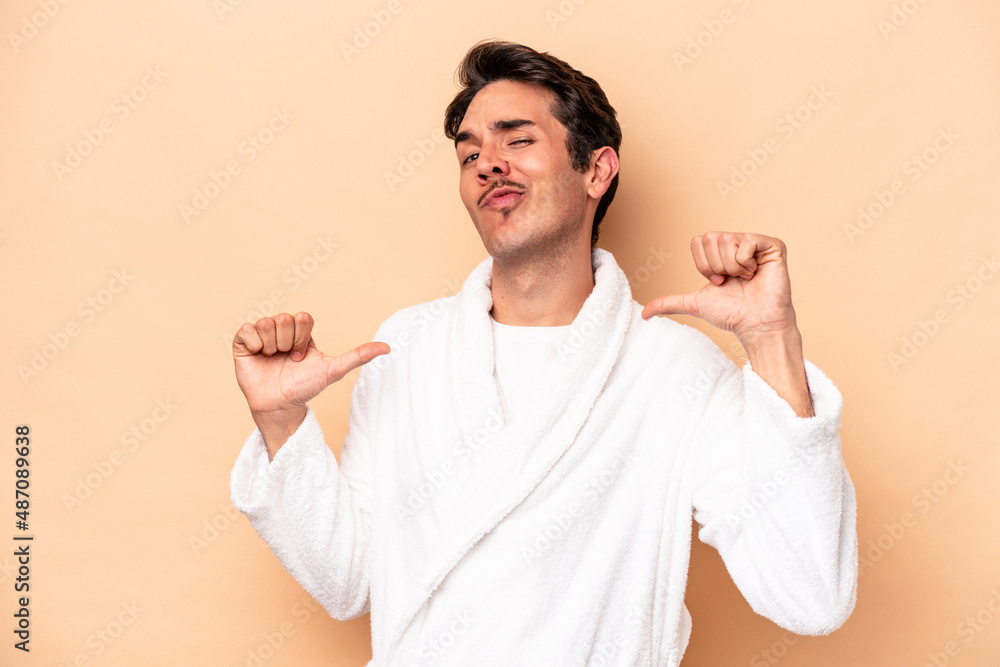 Young caucasian man wearing a bathrobe isolated on beige background feels proud and self confident, example to follow. Young caucasian man wearing a bathrobe isolated on beige background feels proud and self confident, example to follow.
