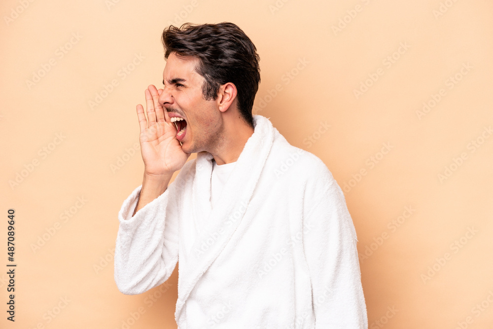 Young caucasian man wearing a bathrobe isolated on beige background shouting and holding palm near opened mouth. Young caucasian man wearing a bathrobe isolated on beige background shouting and holding palm near opened mouth.