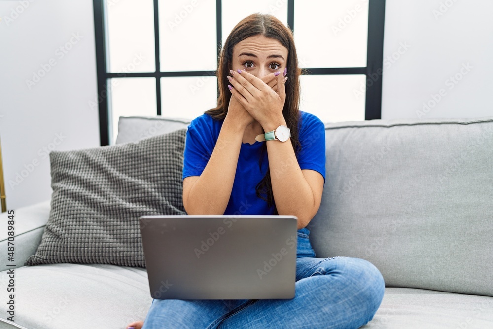 Fototapeta premium Young brunette woman using laptop at home shocked covering mouth with hands for mistake. secret concept.