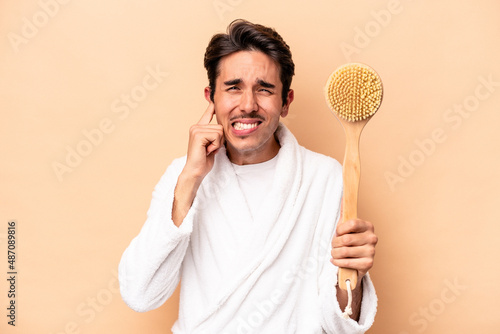 Young caucasian man wearing a bathrobe holding a back scratcher isolated on beige background covering ears with hands.