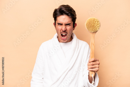 Young caucasian man wearing a bathrobe holding a back scratcher isolated on beige background screaming very angry and aggressive.