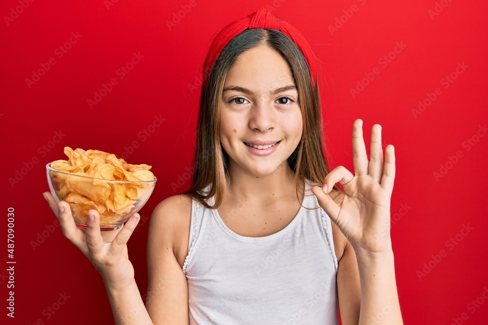 Beautiful brunette little girl holding potato chips doing ok sign with fingers, smiling friendly gesturing excellent symbol Beautiful brunette little girl holding potato chips doing ok sign with fingers, smiling friendly gesturing excellent symbol