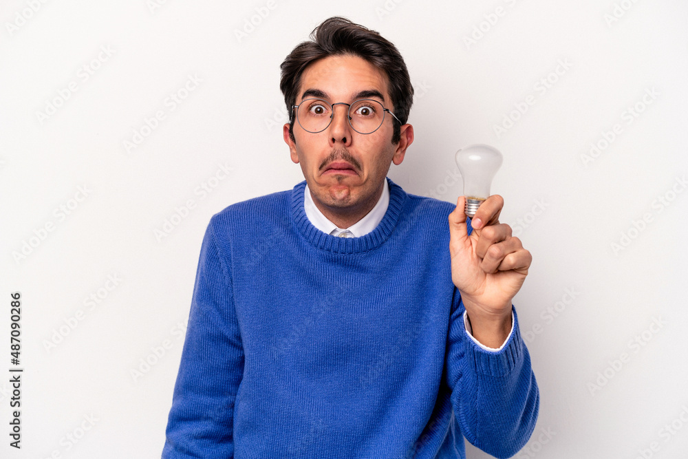 Young caucasian man holding a lightbulb isolated on white background shrugs shoulders and open eyes confused. Young caucasian man holding a lightbulb isolated on white background shrugs shoulders and open eyes confused.
