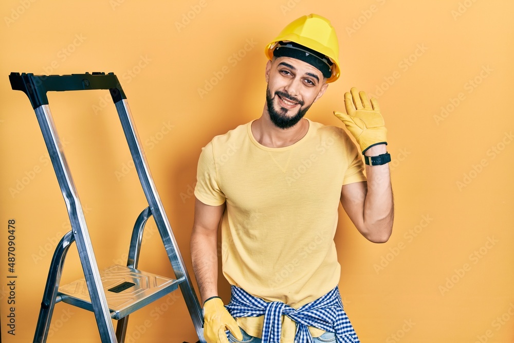 Handsome man with beard by construction stairs wearing hardhat waiving saying hello happy and smiling, friendly welcome gesture Handsome man with beard by construction stairs wearing hardhat waiving saying hello happy and smiling, friendly welcome gesture