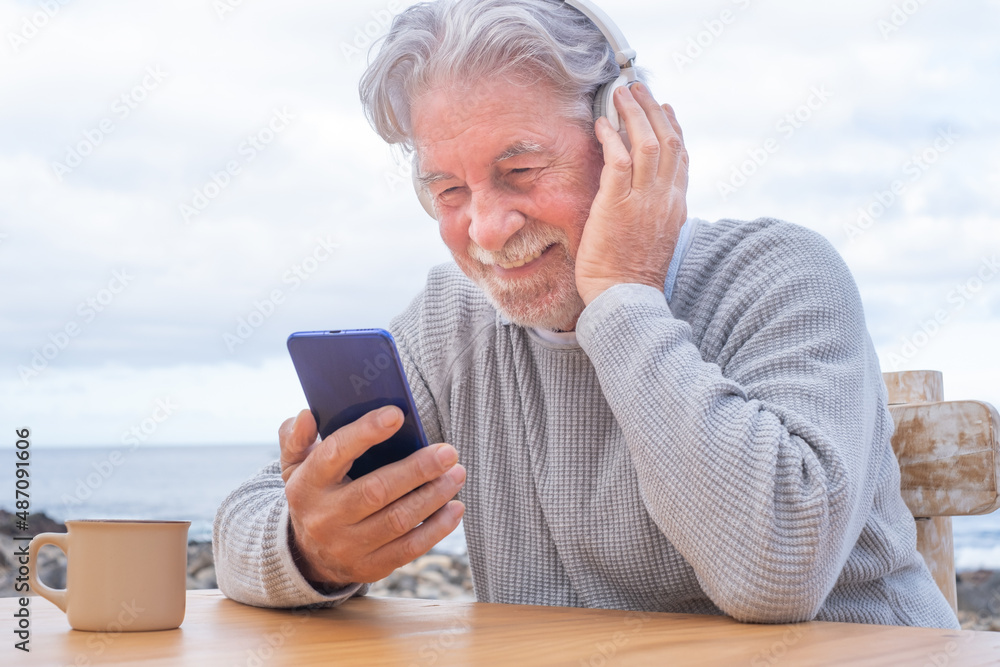 Smiling attractive mature senior adult man with beard using mobile phone listening music with headphones outdoors at sea. White haired elderly man sitting at wooden table enjoying music and coffee cup Smiling attractive mature senior adult man with beard using mobile phone listening music with headphones outdoors at sea. White haired elderly man sitting at wooden table enjoying music and coffee cup