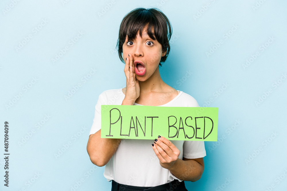 Young hispanic woman holding a plant based placard isolated on blue background is saying a secret hot braking news and looking aside Young hispanic woman holding a plant based placard isolated on blue background is saying a secret hot braking news and looking aside