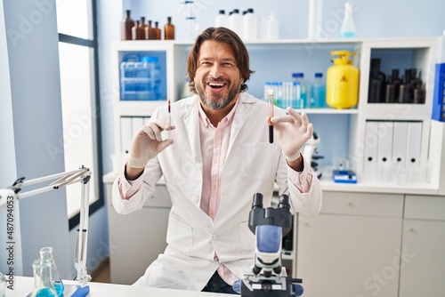 Handsome middle age man holding blood sample at biology laboratory smiling and laughing hard out loud because funny crazy joke.