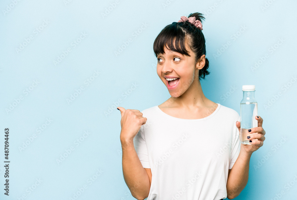 Young hispanic woman holding a bottle of water isolated on blue background points with thumb finger away, laughing and carefree. Young hispanic woman holding a bottle of water isolated on blue background points with thumb finger away, laughing and carefree.