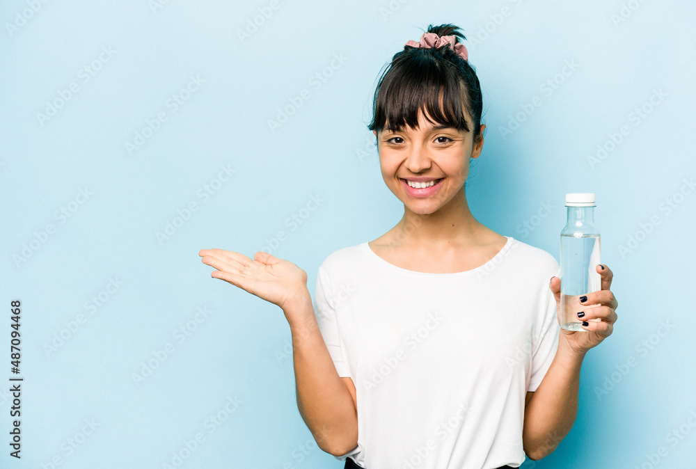 Young hispanic woman holding a bottle of water isolated on blue background showing a copy space on a palm and holding another hand on waist. Young hispanic woman holding a bottle of water isolated on blue background showing a copy space on a palm and holding another hand on waist.