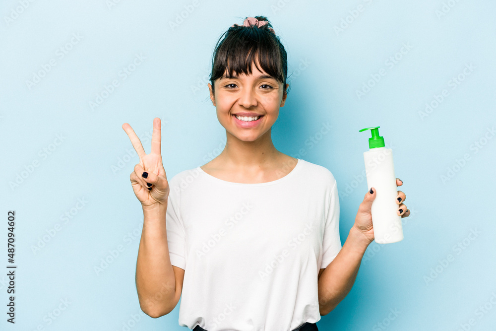 Young hispanic woman holding a body lotion isolated on blue background showing number two with fingers. Young hispanic woman holding a body lotion isolated on blue background showing number two with fingers.