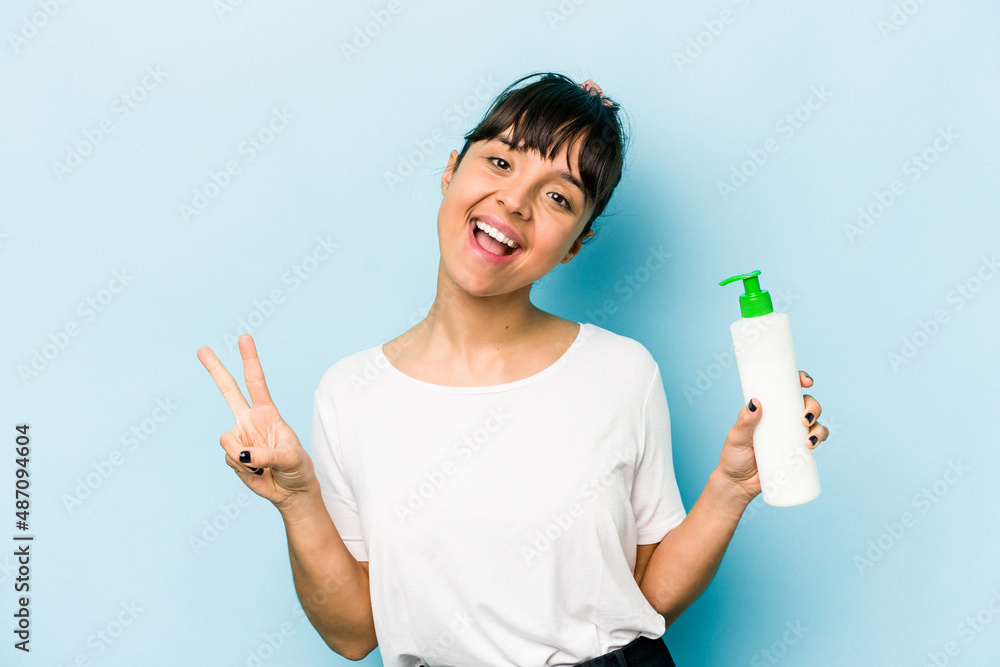 Young hispanic woman holding a body lotion isolated on blue background joyful and carefree showing a peace symbol with fingers. Young hispanic woman holding a body lotion isolated on blue background joyful and carefree showing a peace symbol with fingers.