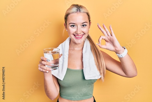 Young blonde girl wearing sportswear drinking glass of water doing ok sign with fingers, smiling friendly gesturing excellent symbol © Krakenimages.com