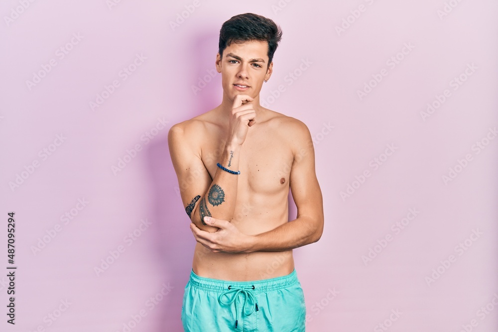 Young hispanic man wearing swimwear shirtless looking confident at the camera with smile with crossed arms and hand raised on chin. thinking positive. Young hispanic man wearing swimwear shirtless looking confident at the camera with smile with crossed arms and hand raised on chin. thinking positive.