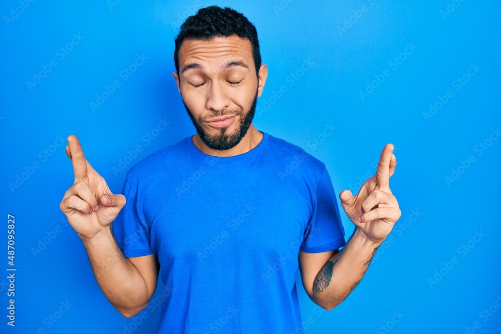Hispanic man with beard wearing casual blue t shirt gesturing finger crossed smiling with hope and eyes closed. luck and superstitious concept. Hispanic man with beard wearing casual blue t shirt gesturing finger crossed smiling with hope and eyes closed. luck and superstitious concept.
