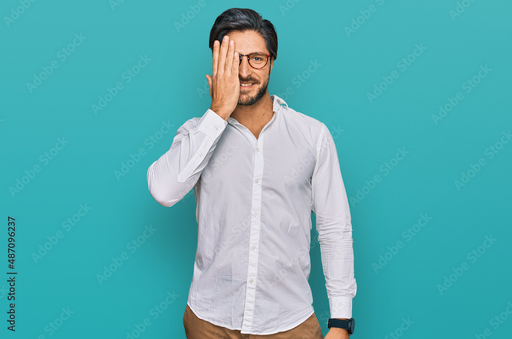 Young hispanic man wearing business shirt and glasses covering one eye with hand, confident smile on face and surprise emotion. Young hispanic man wearing business shirt and glasses covering one eye with hand, confident smile on face and surprise emotion.