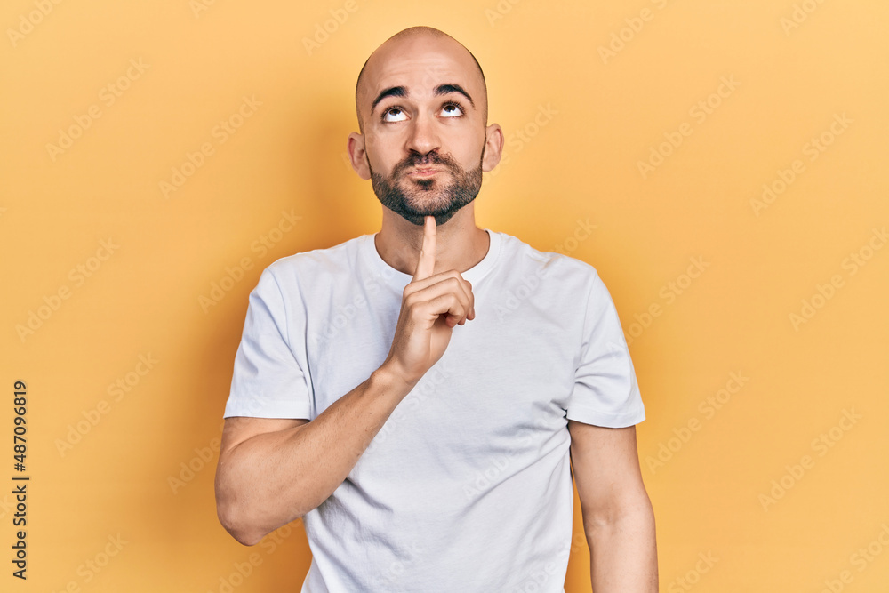 Young bald man wearing casual white t shirt thinking concentrated about doubt with finger on chin and looking up wondering Young bald man wearing casual white t shirt thinking concentrated about doubt with finger on chin and looking up wondering
