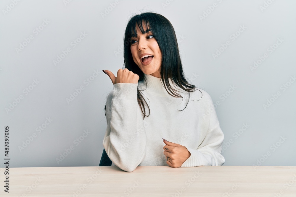 Young brunette woman with bangs wearing casual clothes sitting on the table pointing thumb up to the side smiling happy with open mouth Young brunette woman with bangs wearing casual clothes sitting on the table pointing thumb up to the side smiling happy with open mouth