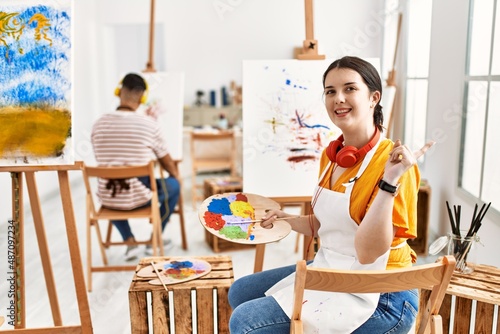 Young artist woman painting on canvas at art studio with a big smile on face, pointing with hand finger to the side looking at the camera. © Krakenimages.com