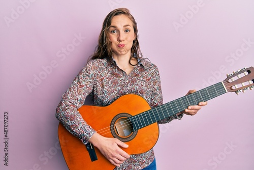 Young blonde woman playing classical guitar puffing cheeks with funny face. mouth inflated with air, catching air.