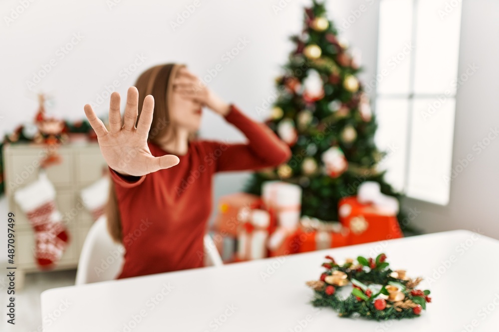 Caucasian young blonde woman sitting on the table by christmas tree covering eyes with hands and doing stop gesture with sad and fear expression. embarrassed and negative concept. Caucasian young blonde woman sitting on the table by christmas tree covering eyes with hands and doing stop gesture with sad and fear expression. embarrassed and negative concept.