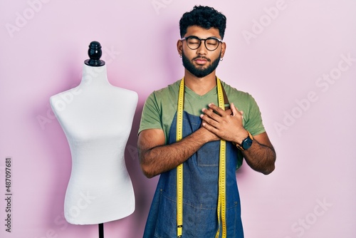 Arab man with beard dressmaker designer wearing atelier apron smiling with hands on chest, eyes closed with grateful gesture on face. health concept.