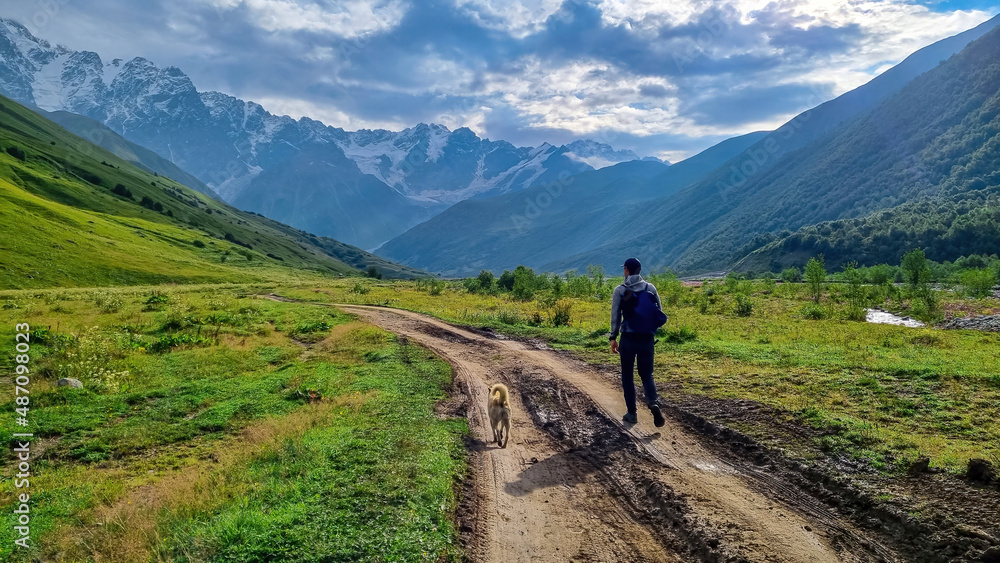 A man walking with a dog toward the Shkhara Glacier in the Greater Caucasus Mountain Range in Georgia, Svaneti Region, Ushguli. Stray dog. Wanderlust, hiking. Humans best friend. Outdoor activities.