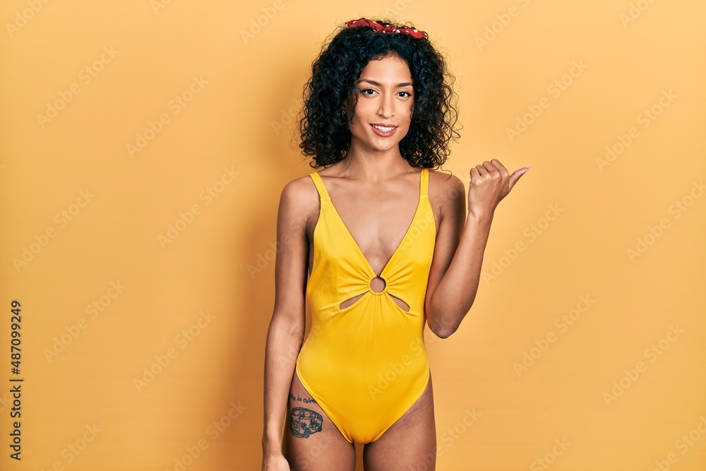 Young latin girl wearing swimsuit smiling with happy face looking and pointing to the side with thumb up. Young latin girl wearing swimsuit smiling with happy face looking and pointing to the side with thumb up.