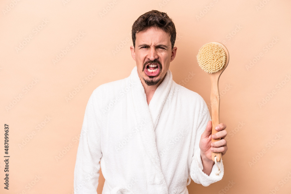 Young caucasian man holding back scratcher isolated on beige background screaming very angry and aggressive. Young caucasian man holding back scratcher isolated on beige background screaming very angry and aggressive.